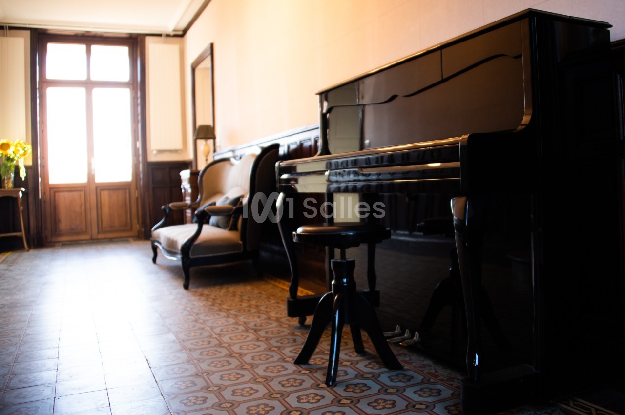 Piano droit noir dans un salon lumineux avec carrelage ancien, fauteuils en bois et une fenêtre en arrière-plan.