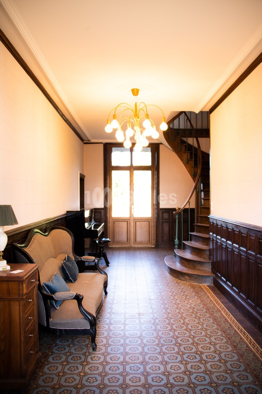 Couloir lumineux avec carrelage ancien, canapé, piano, escalier en colimaçon et porte-fenêtre en arrière-plan.