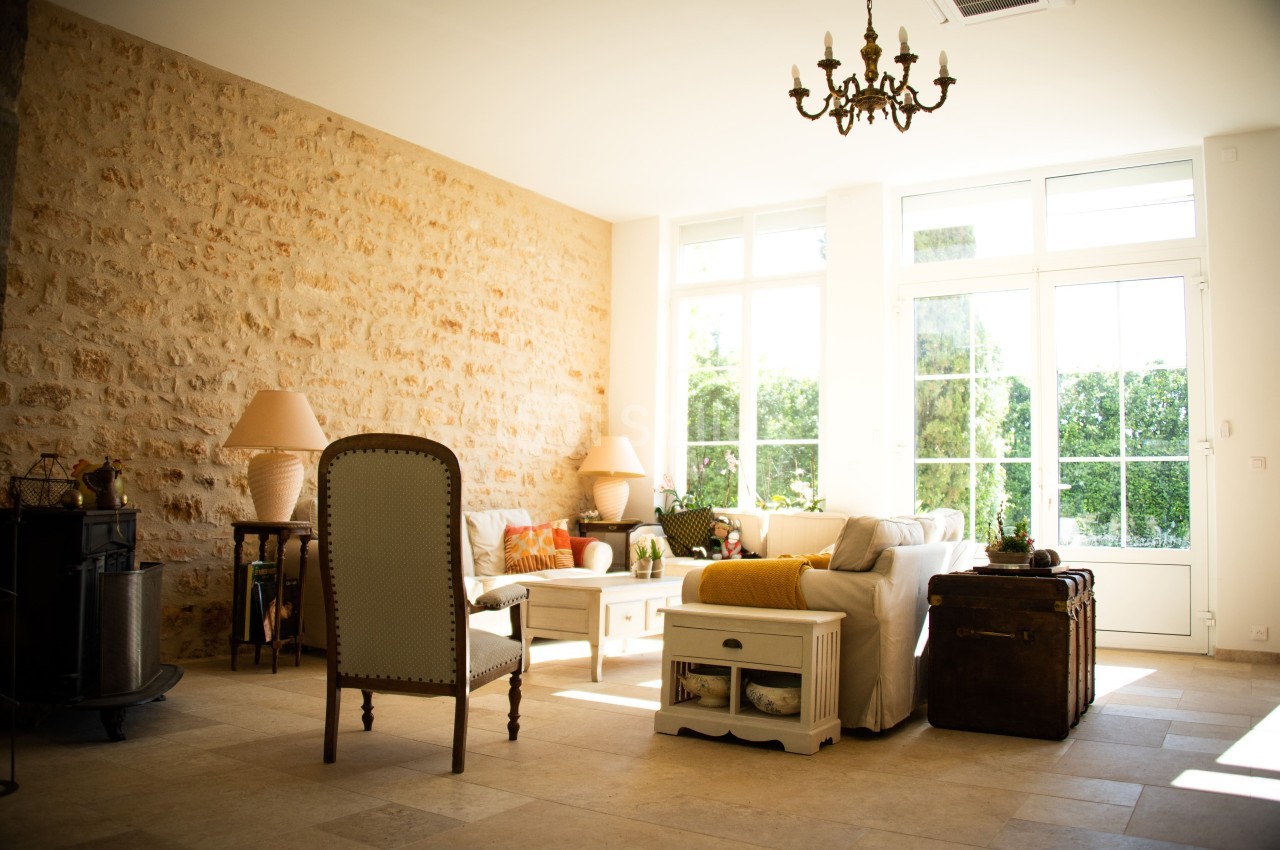 Salon lumineux avec mur en pierre, canapé beige, fauteuil, table basse et grandes fenêtres donnant sur un jardin.
