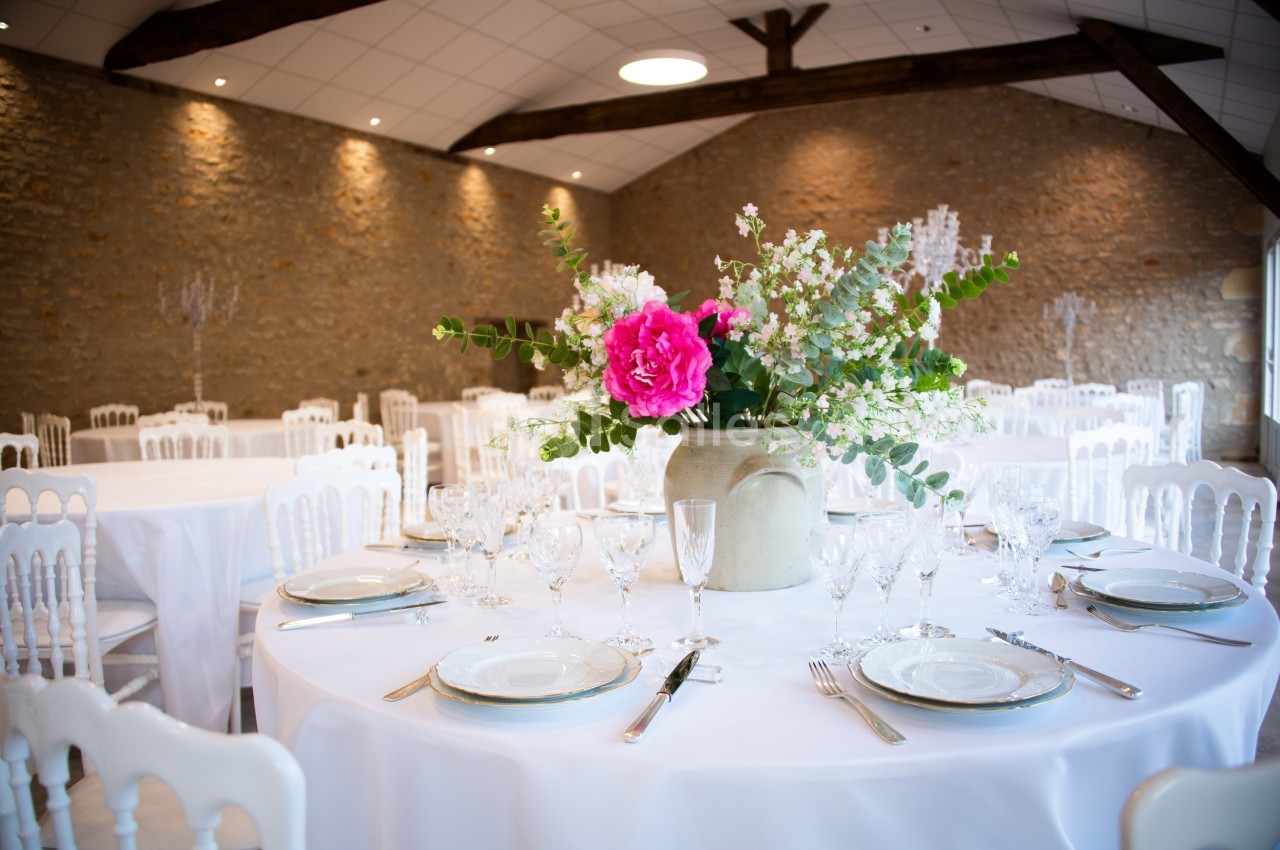 Table ronde décorée avec une nappe blanche, vaisselle élégante et un bouquet de fleurs au centre, dans une salle en pierre.