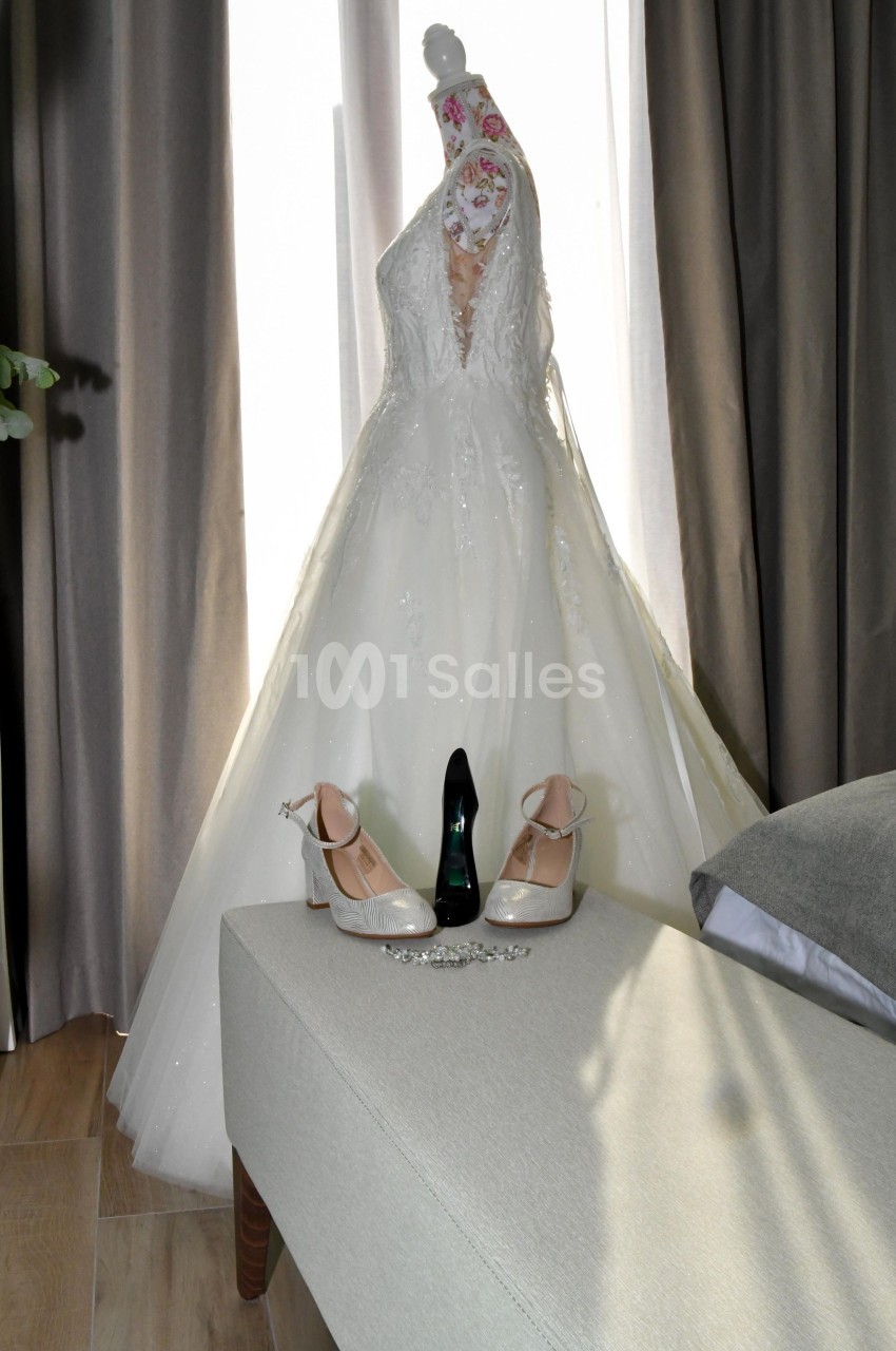 Robe de mariée blanche sur un mannequin, entourée de chaussures, d'un bijou et d'un rideau en arrière-plan.