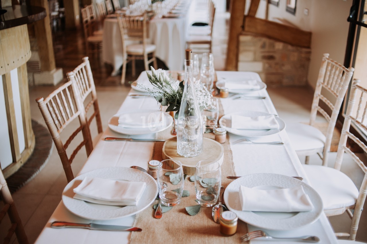 Table dressée avec nappes blanches, assiettes, verres, couverts et décoration florale dans une salle lumineuse.