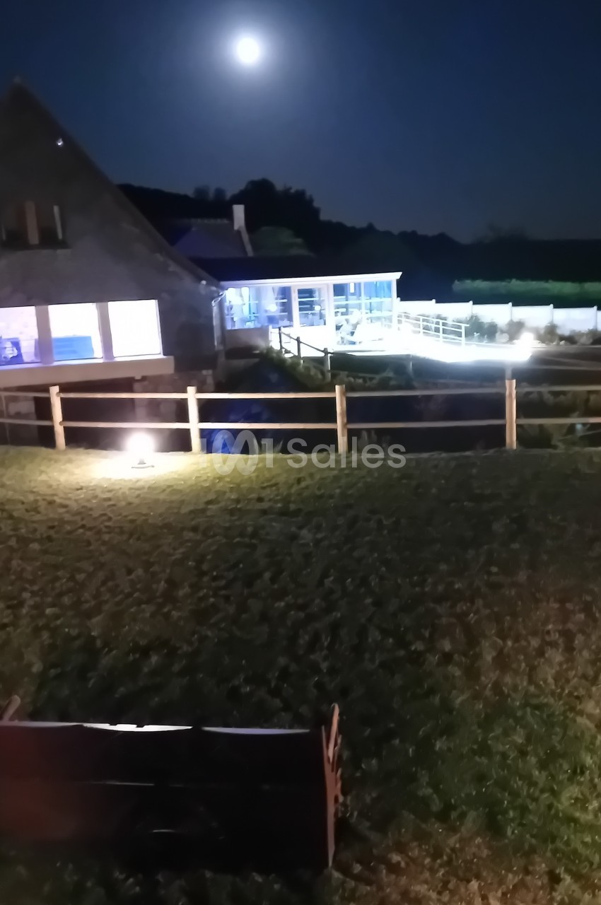 Vue nocturne d'un jardin avec une maison éclairée, une clôture en bois, un banc au premier plan et la lune visible.