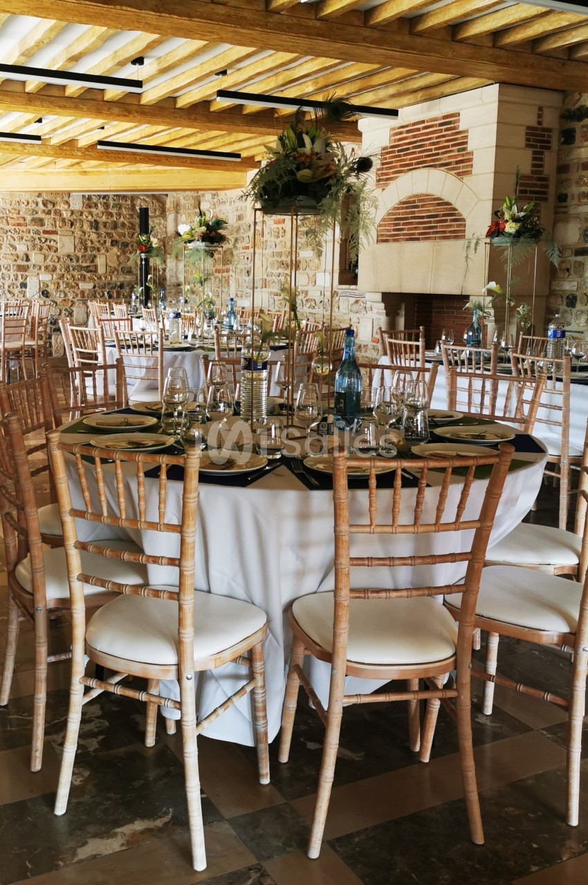 Salle de réception avec tables rondes dressées, chaises en bois clair et décorations florales élégantes.