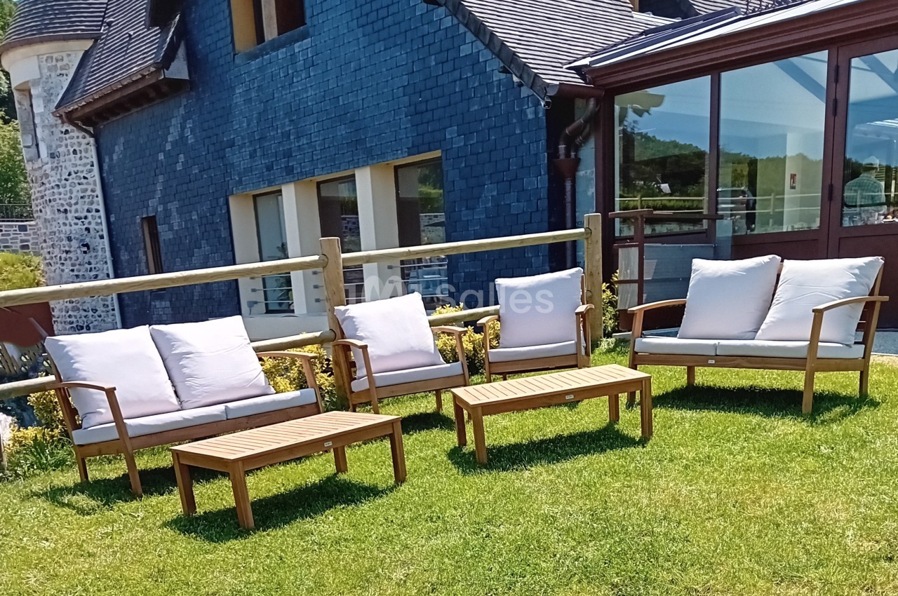 Mobilier de jardin en bois avec coussins blancs disposé sur une pelouse devant une maison en ardoise.