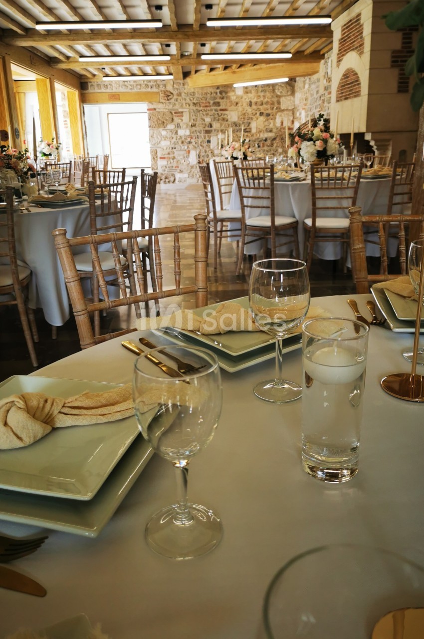 Salle de réception avec tables dressées, nappes blanches, verres, assiettes et décorations florales dans un cadre rustique.