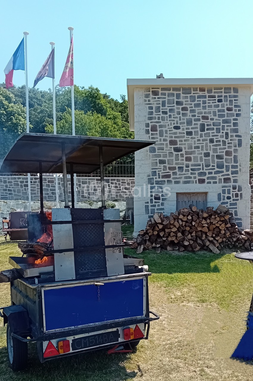 Barbecue mobile en métal allumé devant un bâtiment en pierre, avec des drapeaux et un tas de bois à proximité.