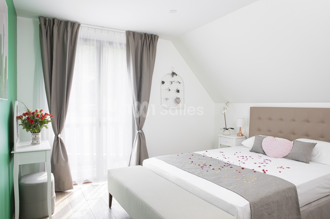 Chambre lumineuse avec lit double, draps décorés de pétales, rideaux gris, bureau avec vase de fleurs et lumière naturelle.