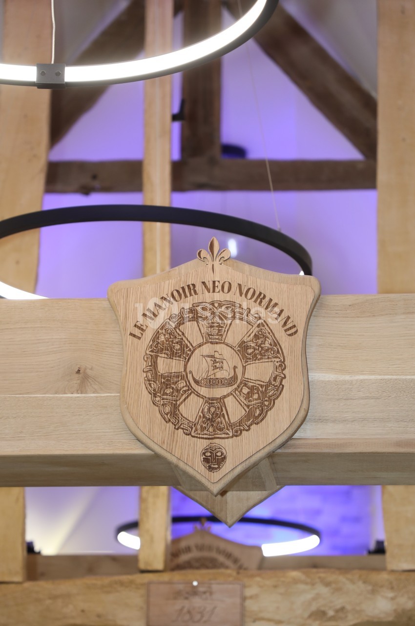 Blason en bois gravé ’Le Manoir Néo-Normand’ suspendu dans un intérieur avec poutres apparentes et éclairage moderne.