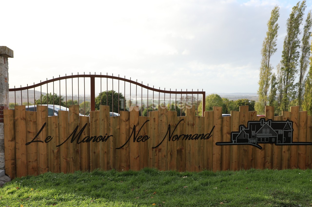 Clôture en bois avec le texte ’Le Manoir Néo Normand’ et un dessin de maison, devant un paysage rural.
