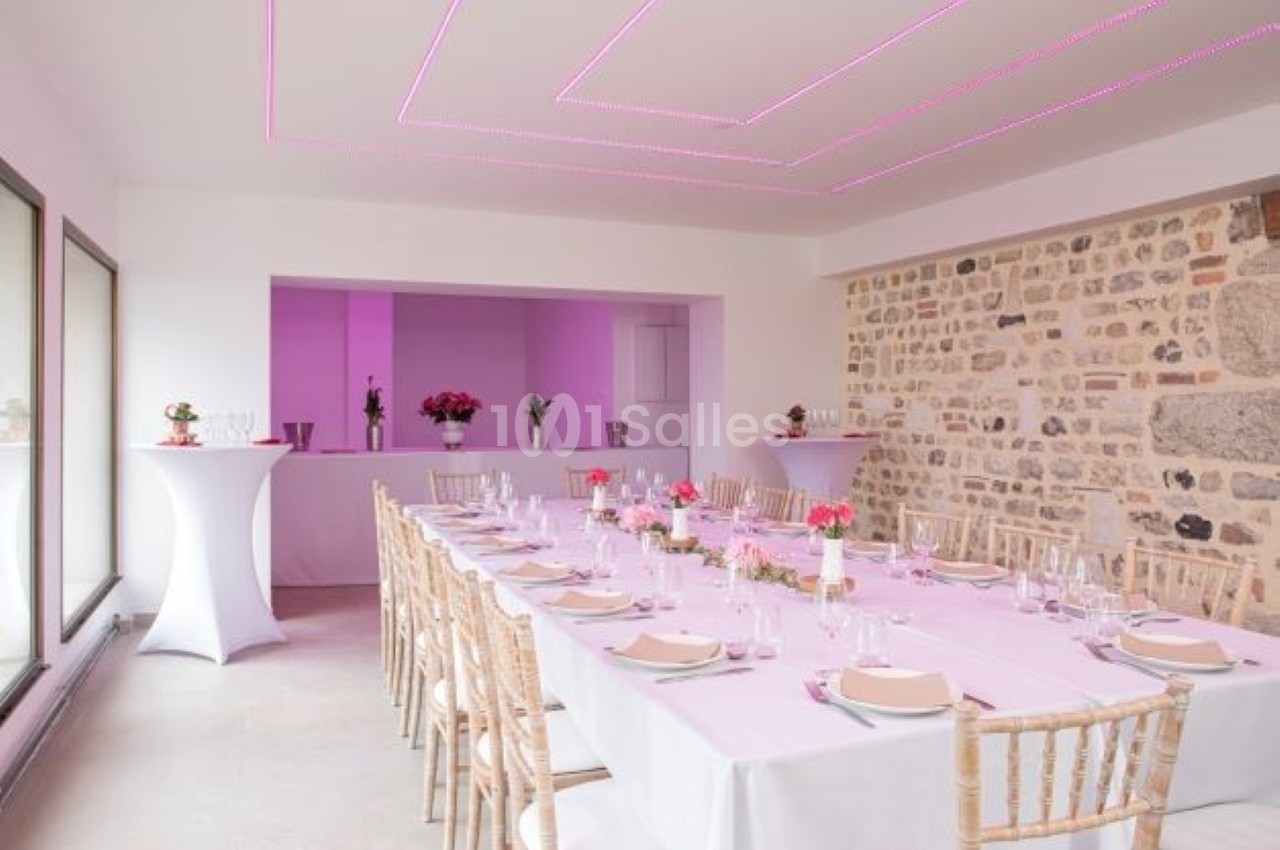 Salle de réception lumineuse avec une longue table dressée, décorée de fleurs roses et éclairée par des lumières violettes.
