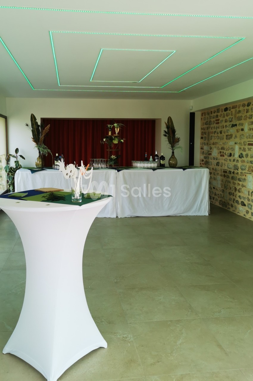 Salle de réception avec table haute en premier plan et buffet décoré de plantes et nappes blanches en arrière-plan.