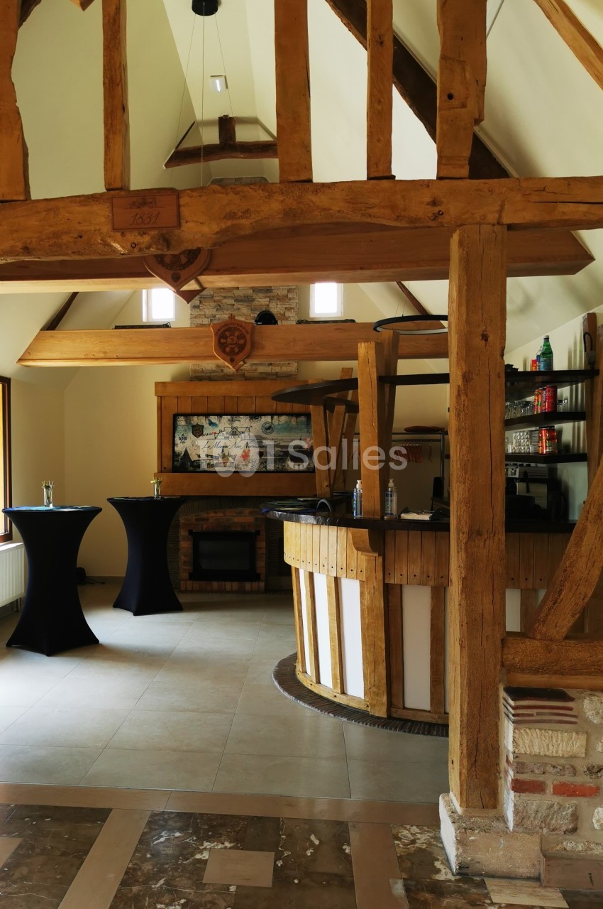 Intérieur d'une salle avec poutres apparentes, bar en bois arrondi et tables hautes habillées de nappes noires.