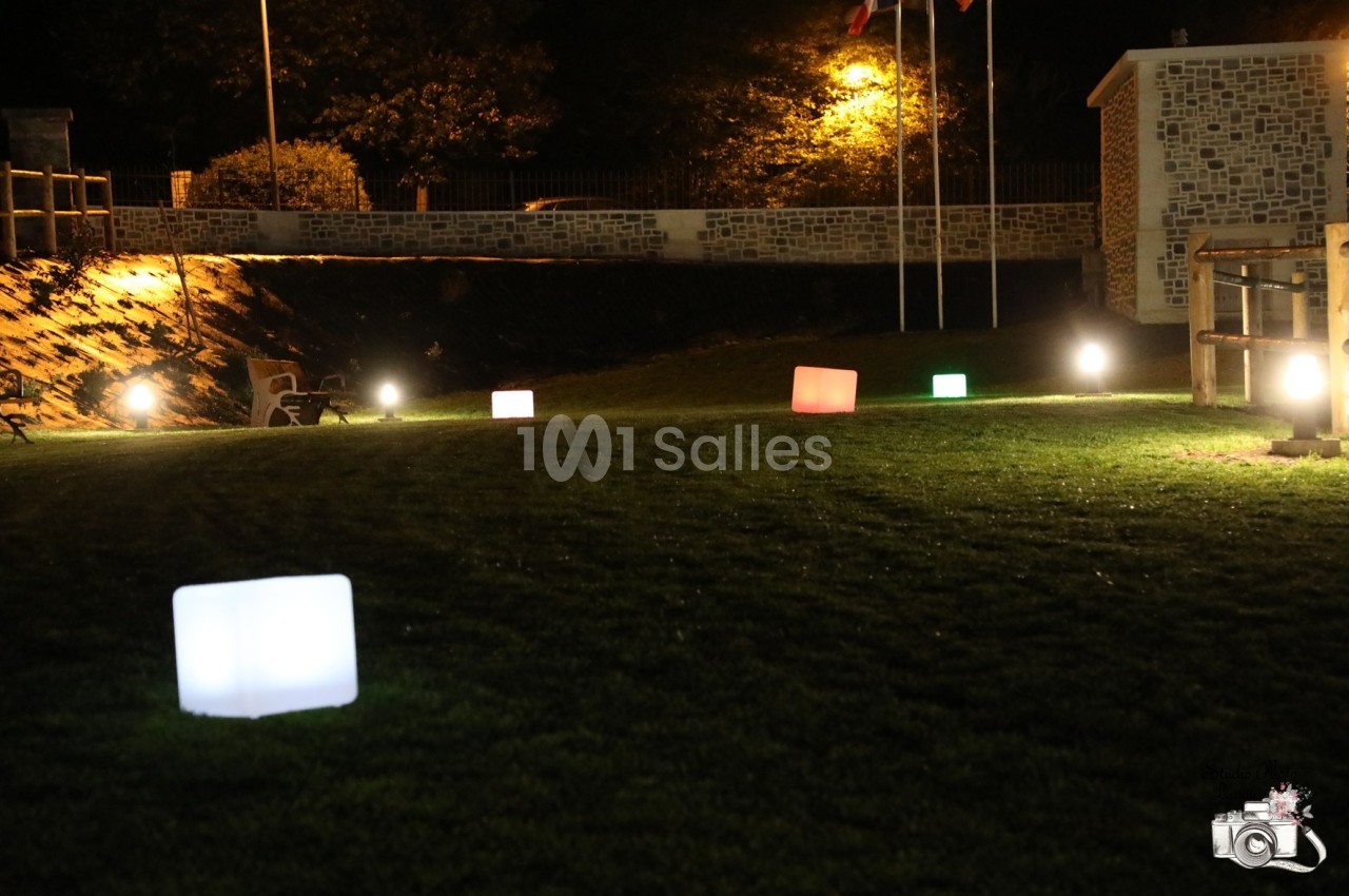 Carré de pelouse éclairé par des cubes lumineux colorés et des lampadaires, avec un mur en pierre en arrière-plan.