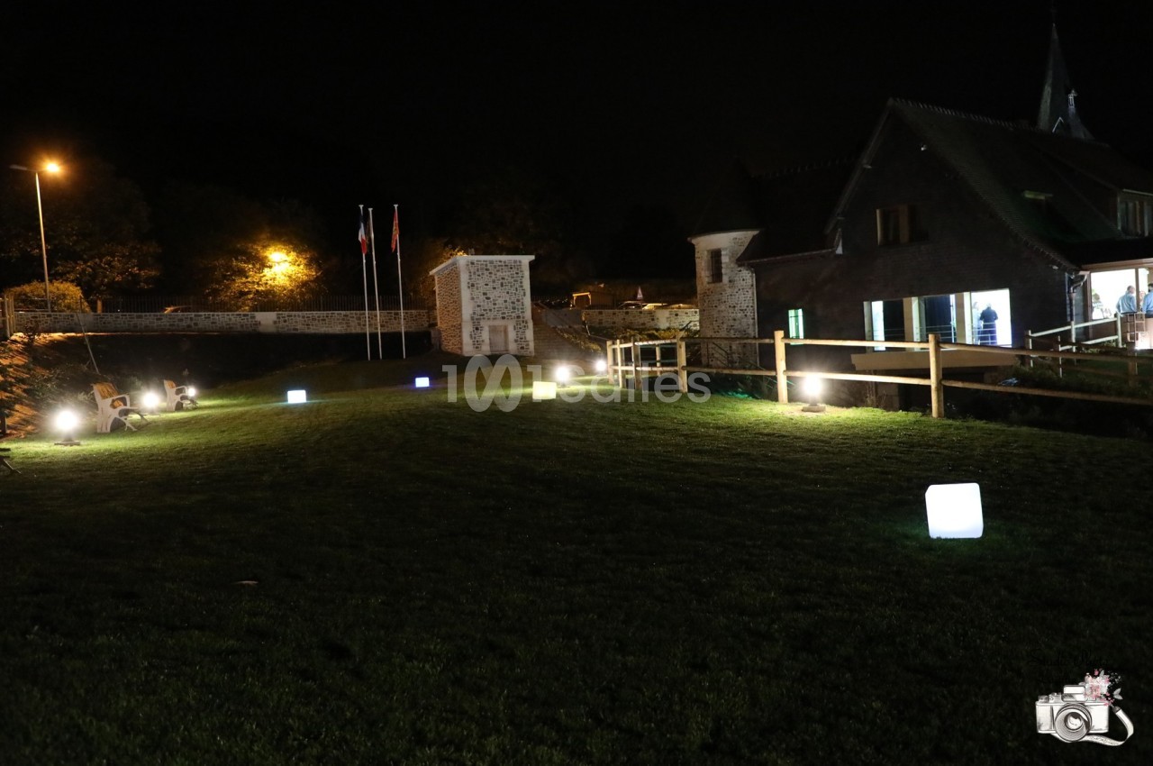 Pelouse éclairée par des cubes lumineux colorés, avec un bâtiment en arrière-plan et un pont en bois.