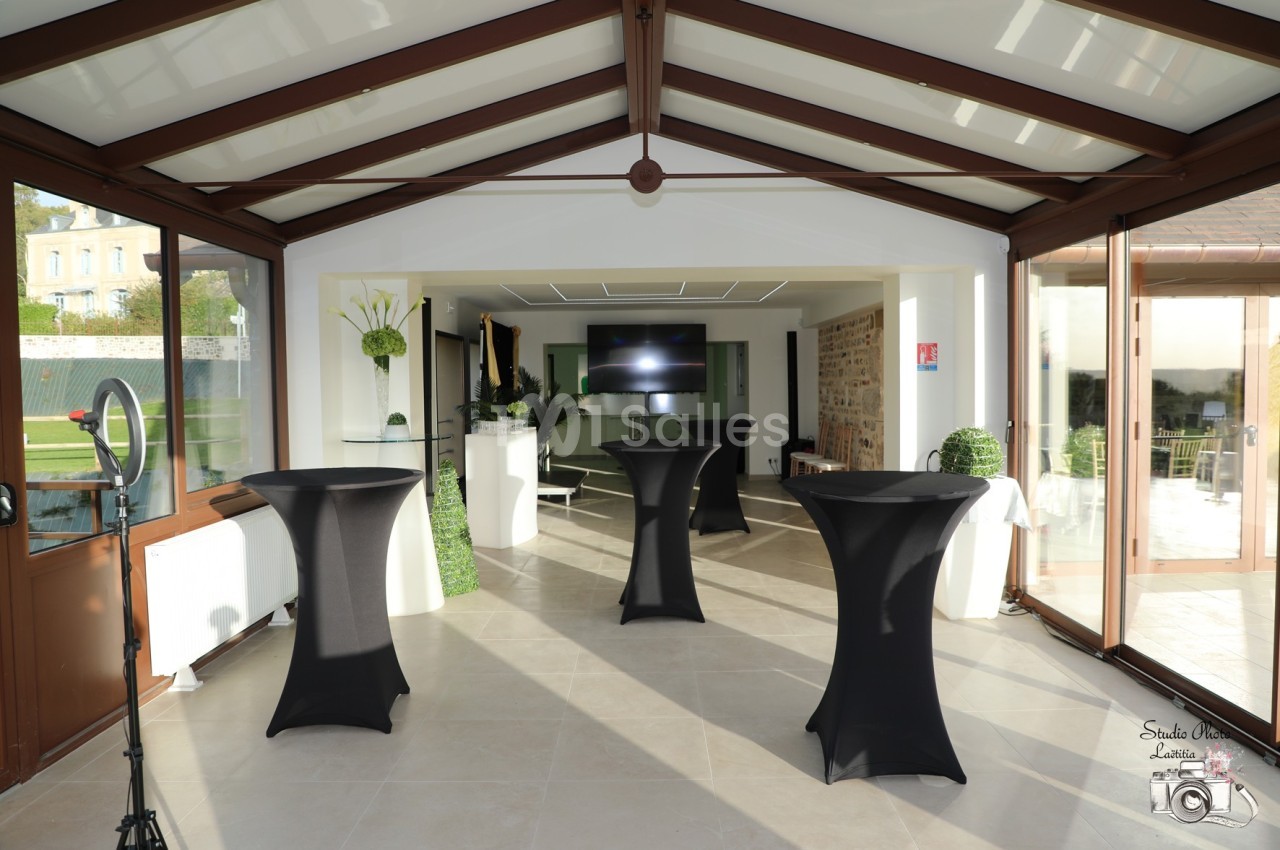 Salle lumineuse avec tables hautes noires, grandes baies vitrées, écran au fond et vue sur un jardin.