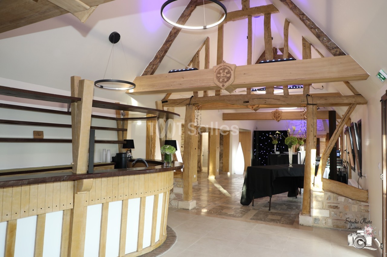 Intérieur d'une salle avec charpente en bois apparente, comptoir en bois et tables décorées pour un événement.