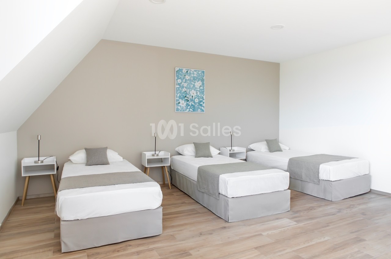 Trois lits simples alignés dans une chambre lumineuse avec parquet et mur beige décoré d'un tableau floral.