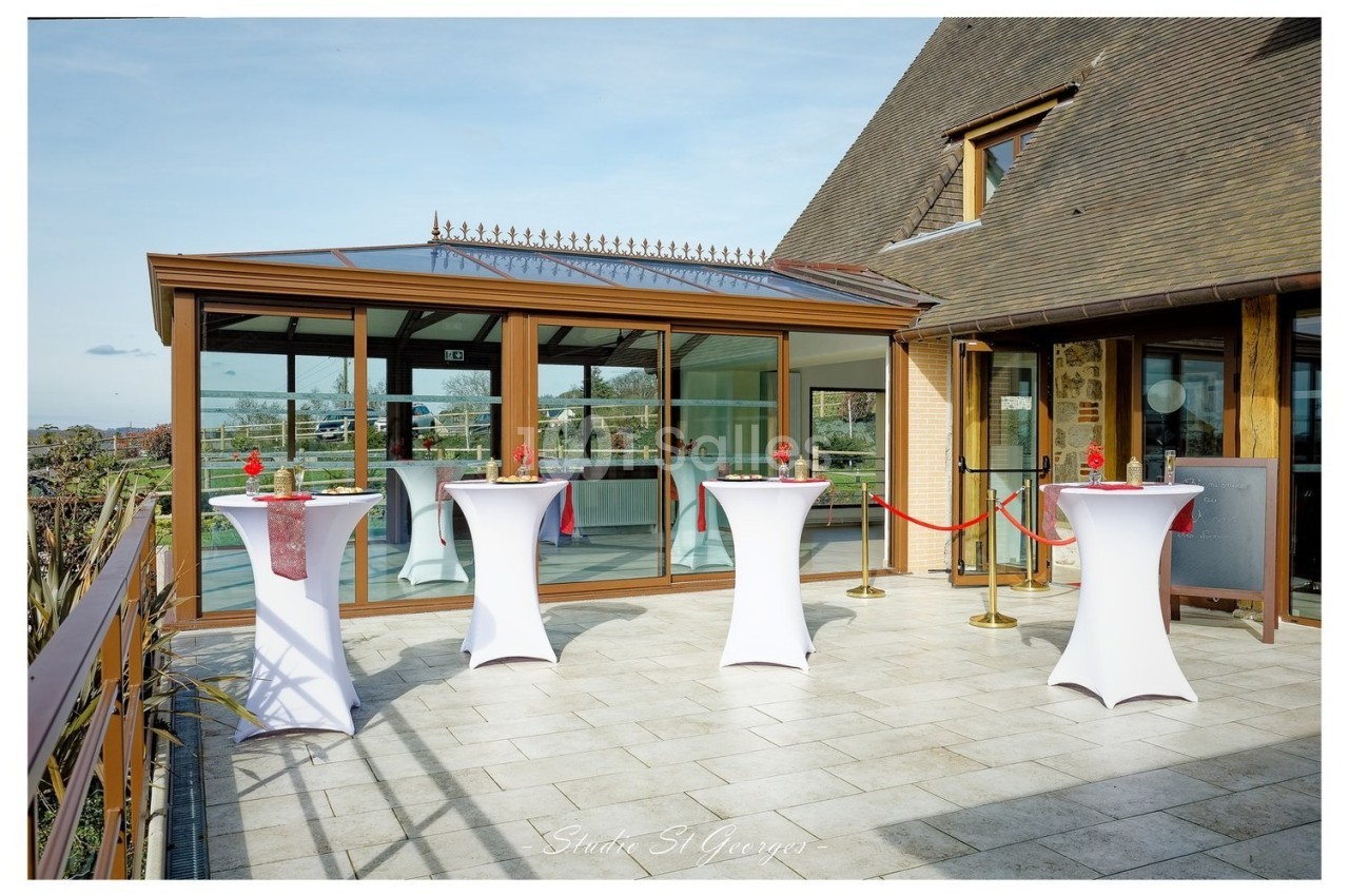 Terrasse avec tables hautes drapées de blanc, décorées de fleurs rouges, devant une salle vitrée.