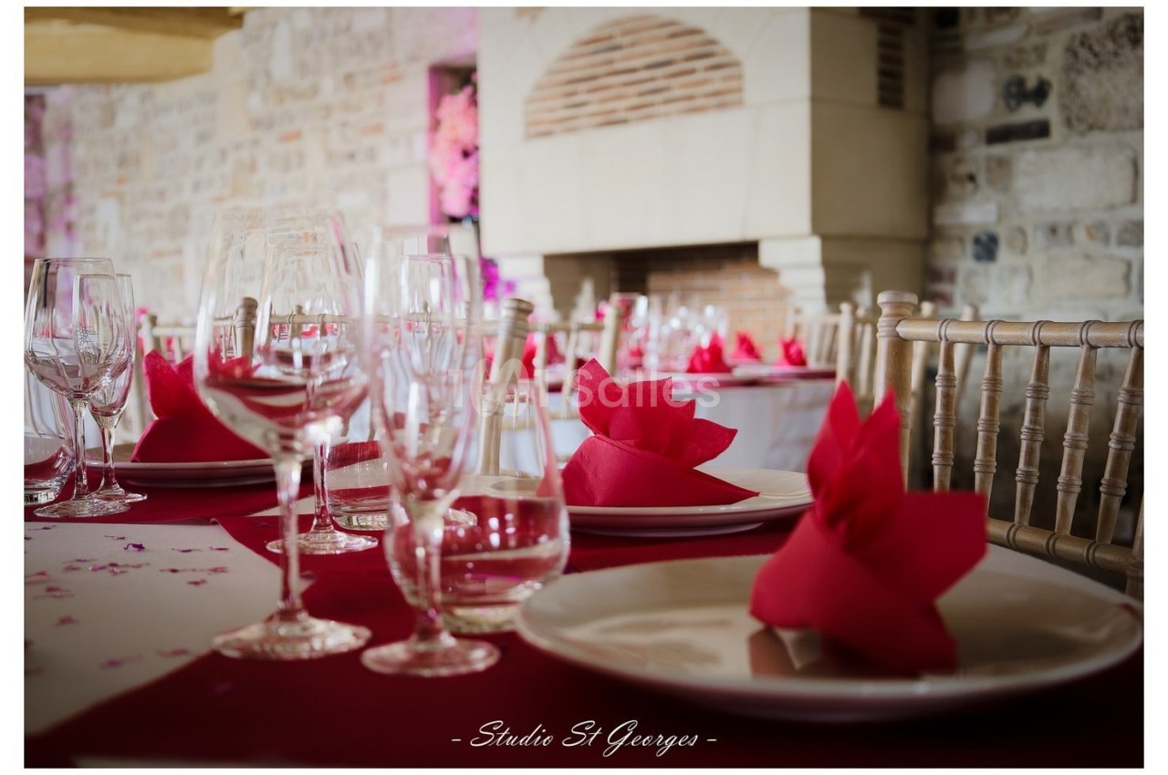 Table dressée avec nappes rouges, serviettes pliées en éventail, verres et assiettes, devant une cheminée en pierre.
