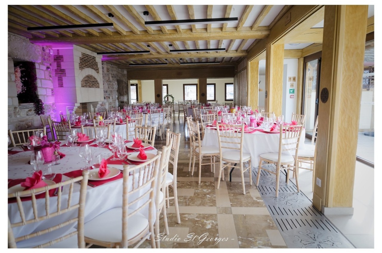Salle de réception lumineuse avec tables rondes décorées de nappes blanches et serviettes roses, chaises en bois clair.