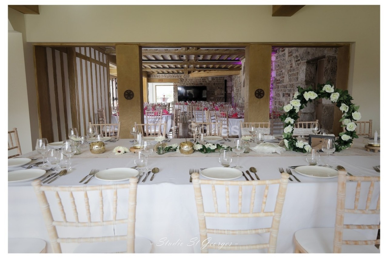 Table de réception élégamment dressée avec des chaises en bois clair, décorations florales et vue sur une salle en pierre.