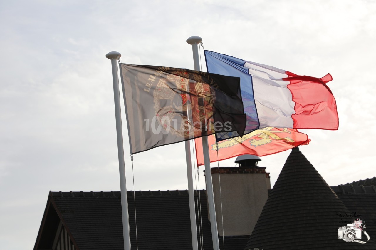 Trois drapeaux flottant au vent, dont le drapeau français, devant des toits en ardoise et une cheminée.