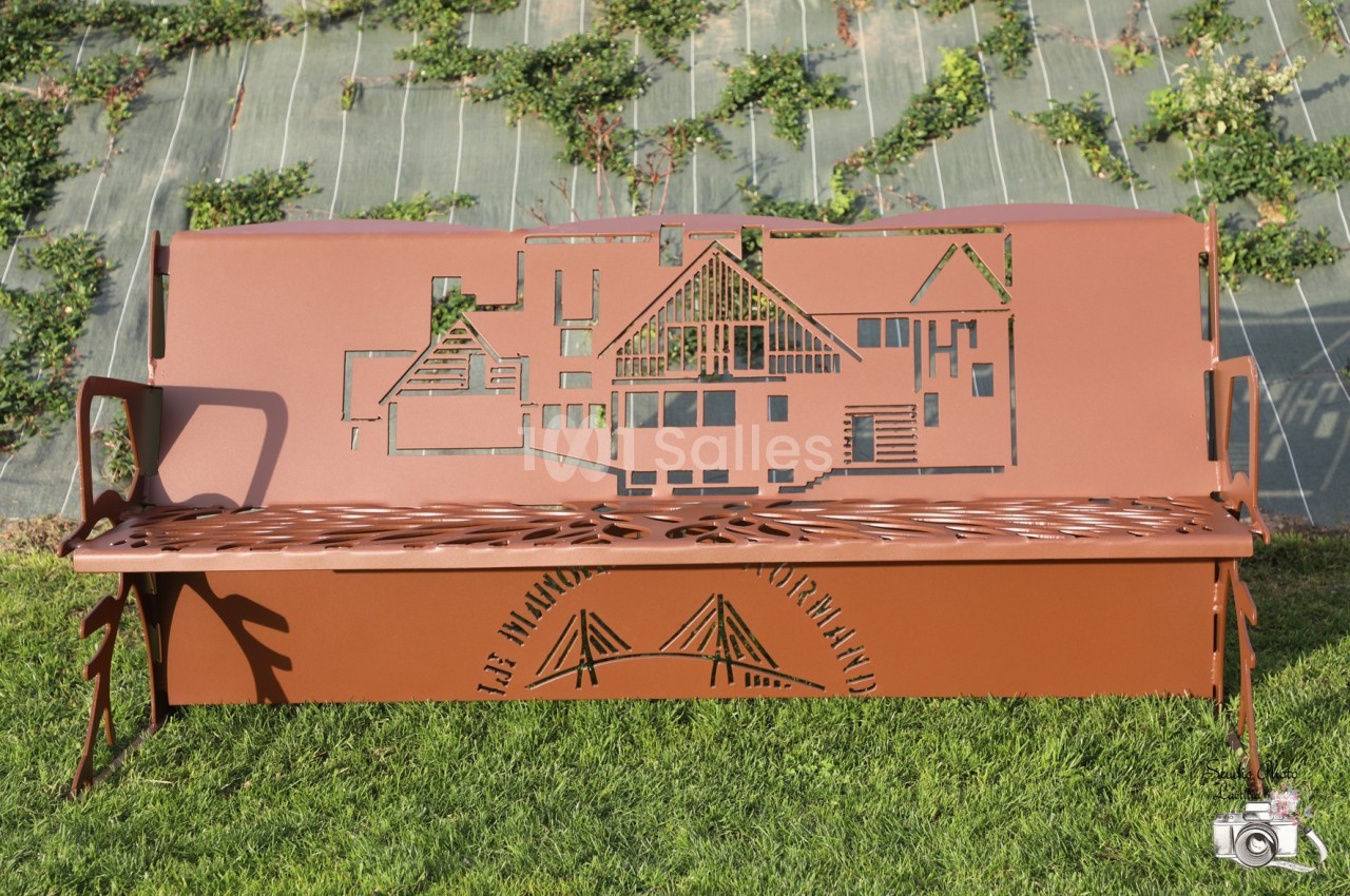 Banc en métal rouillé avec des motifs découpés représentant des bâtiments et un pont, installé sur une pelouse.