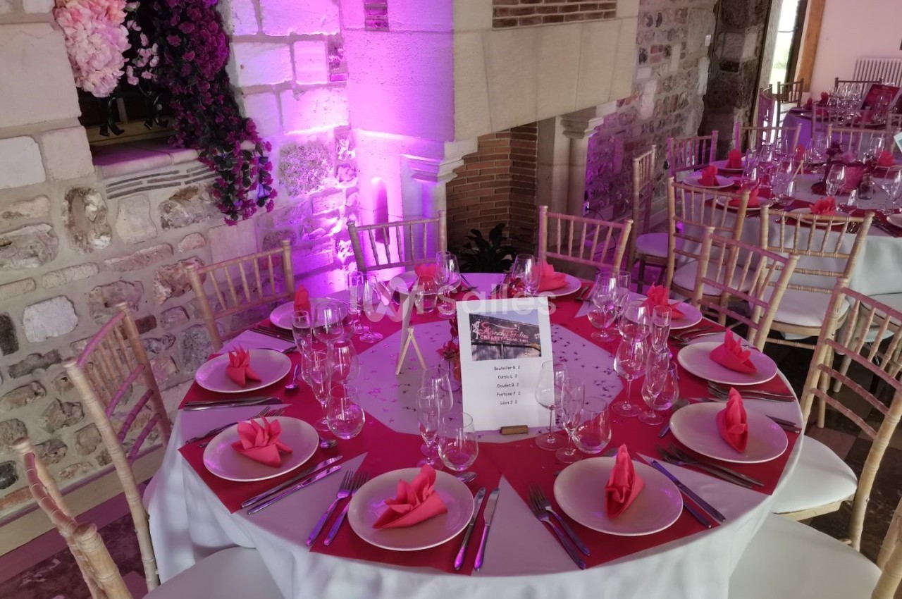 Table ronde décorée pour un événement, avec nappes roses, serviettes pliées, verres et couverts, dans une salle en pierre.