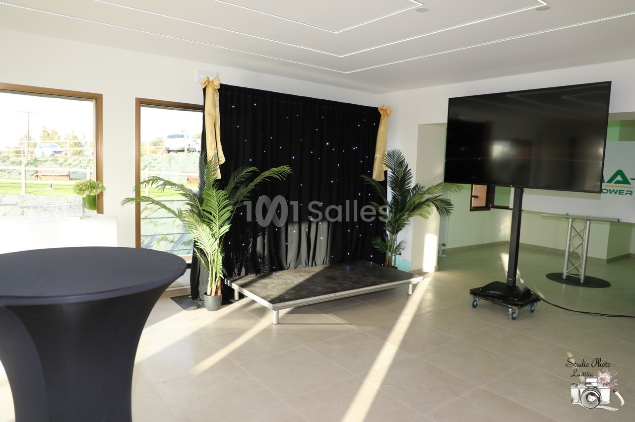 Espace intérieur lumineux avec une petite scène, rideau noir étoilé, écran sur pied et plantes décoratives.