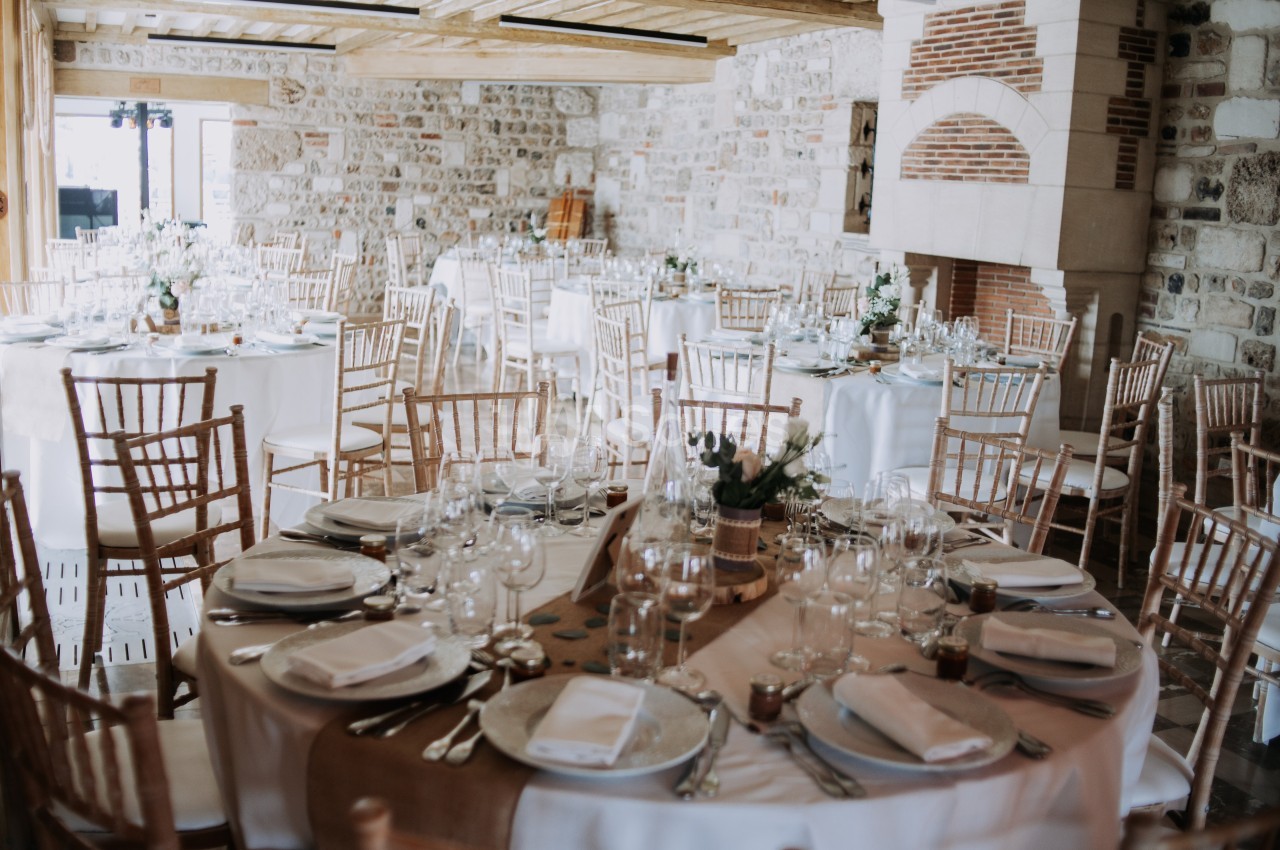 Salle de réception lumineuse avec des tables rondes décorées pour un événement, entourées de chaises en bois.