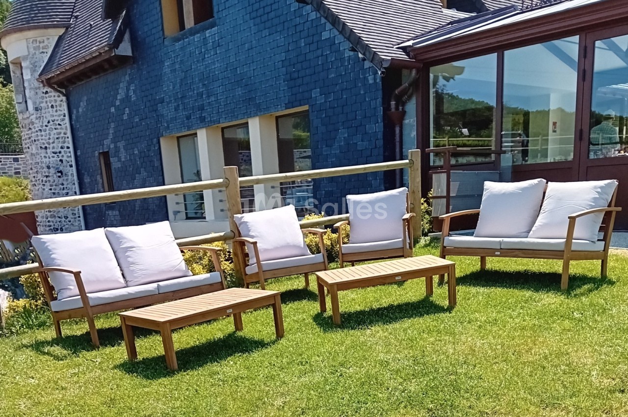 Mobilier de jardin en bois avec coussins blancs disposé sur une pelouse devant une maison en ardoise.