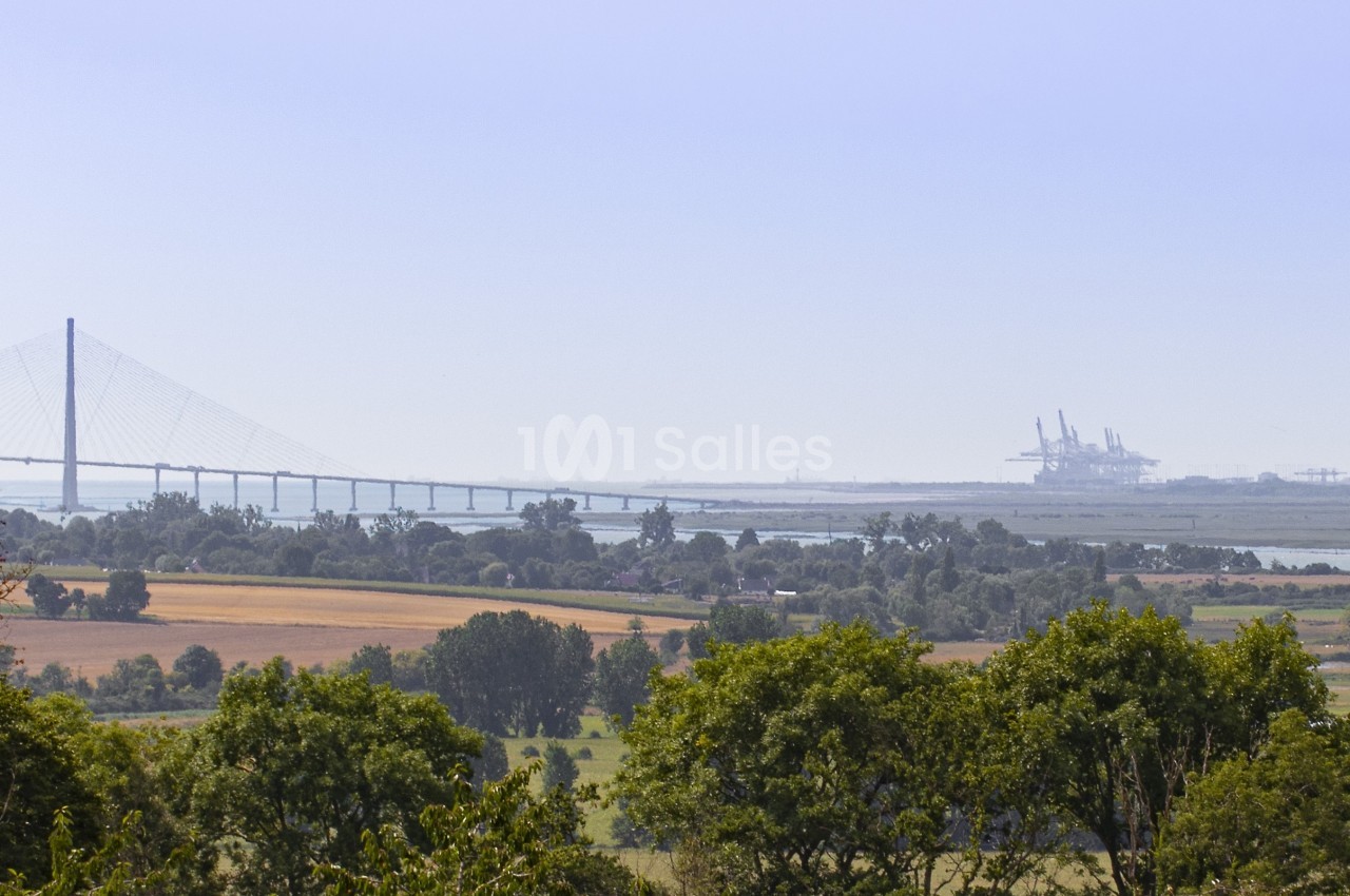 Paysage rural avec arbres, champs et vue lointaine sur un pont suspendu et des installations industrielles.