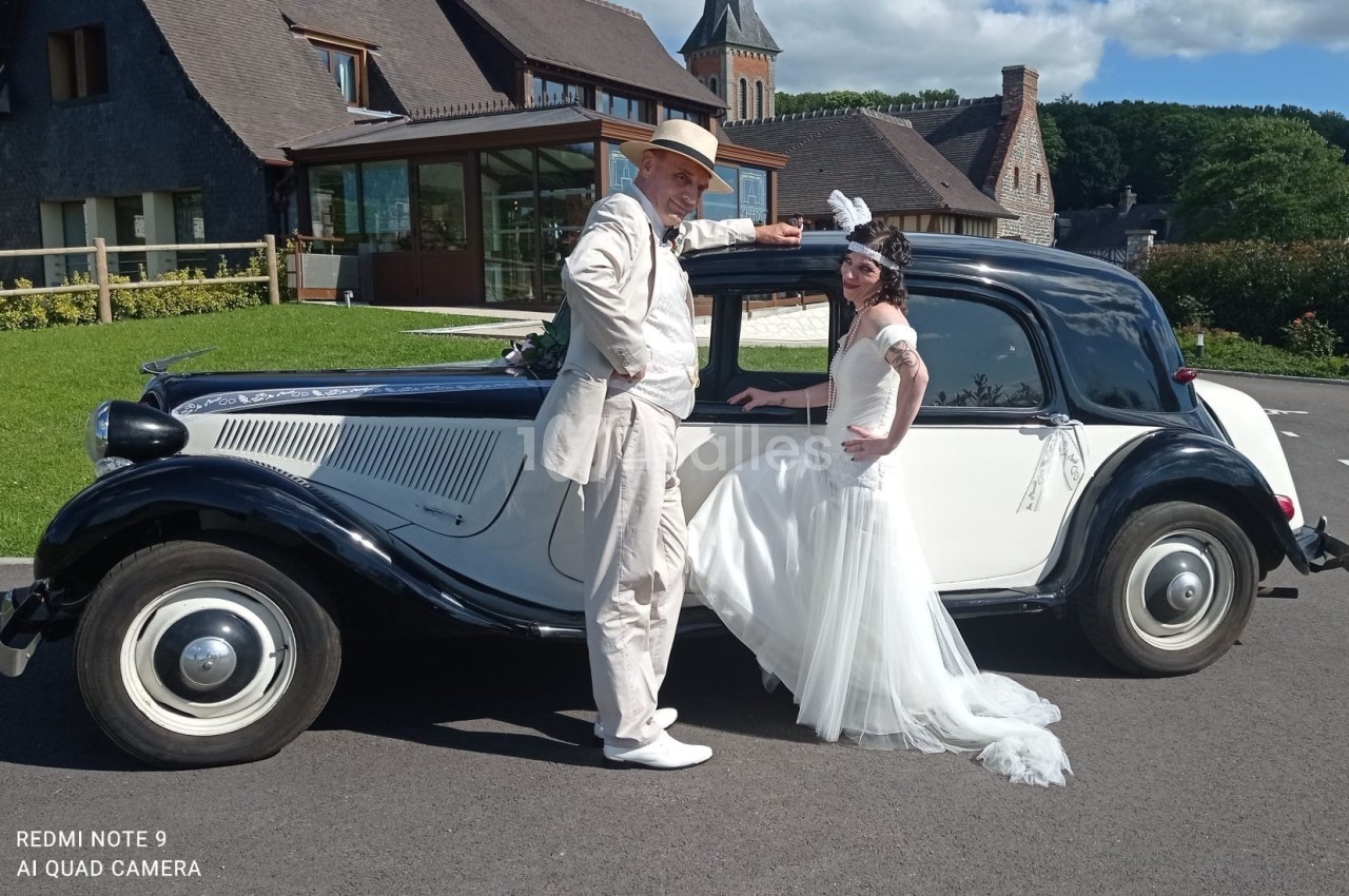 Un couple en tenue de mariage pose devant une voiture ancienne noire et blanche, sur une route près d'un bâtiment.