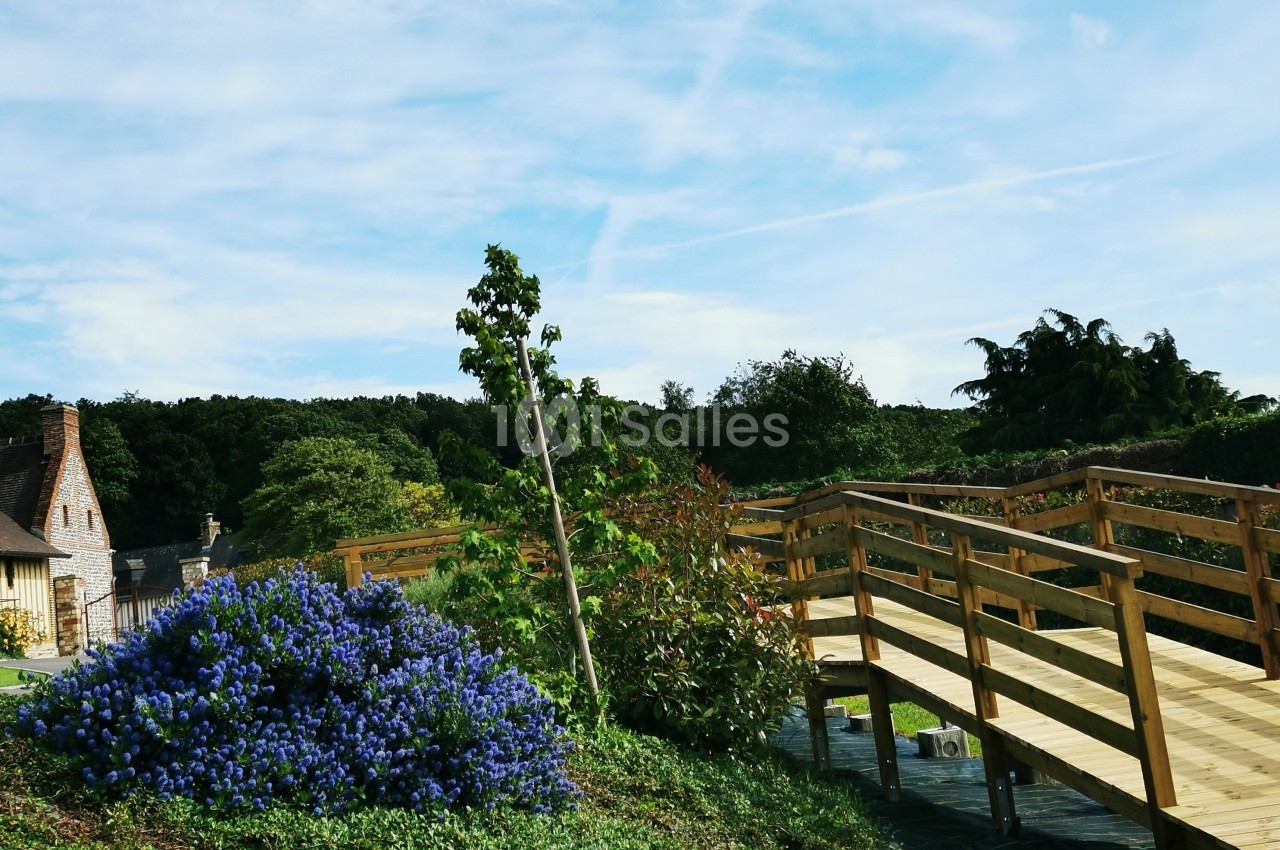 Passerelle en bois traversant un jardin verdoyant avec des fleurs bleues et une maison en arrière-plan.