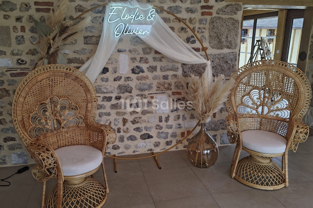 Deux fauteuils en rotin devant un mur en pierre, décorés avec un cercle orné de voilages et de pampas.