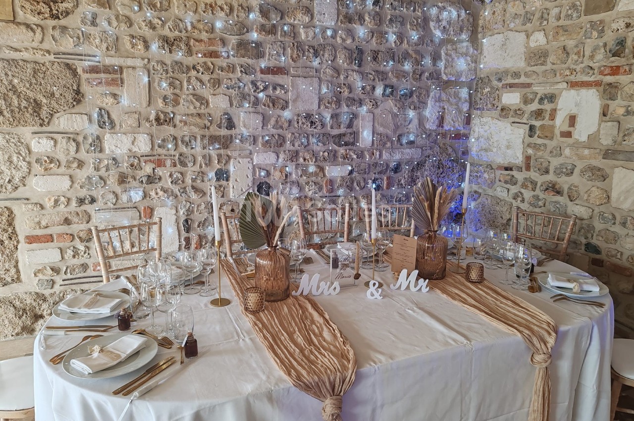 Table décorée pour un mariage avec nappes beige, vaisselle élégante, bougies et ornements devant un mur en pierre.