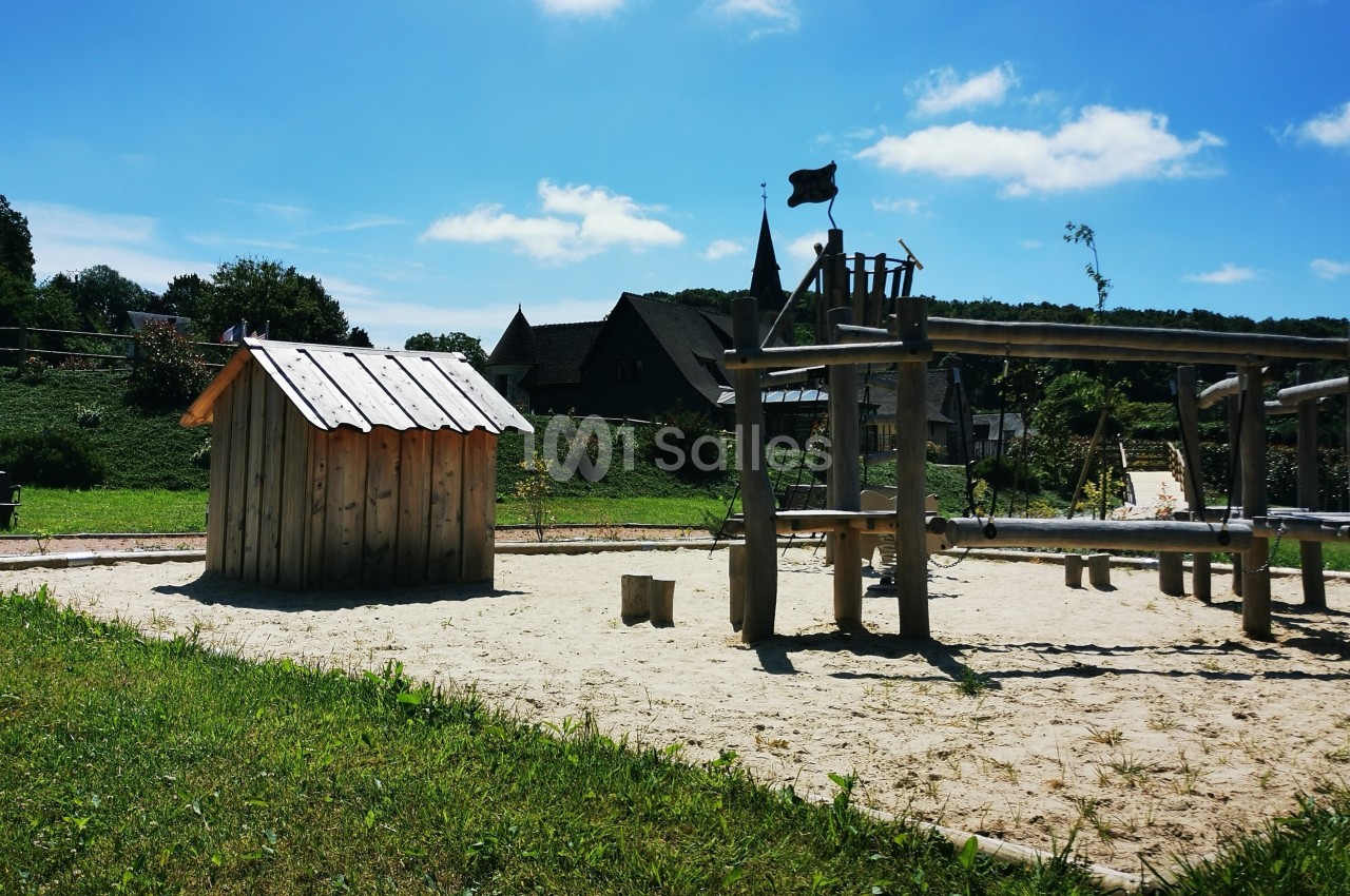 Aire de jeux en bois sur sol sablé, entourée de verdure et de bâtiments en arrière-plan sous un ciel bleu.