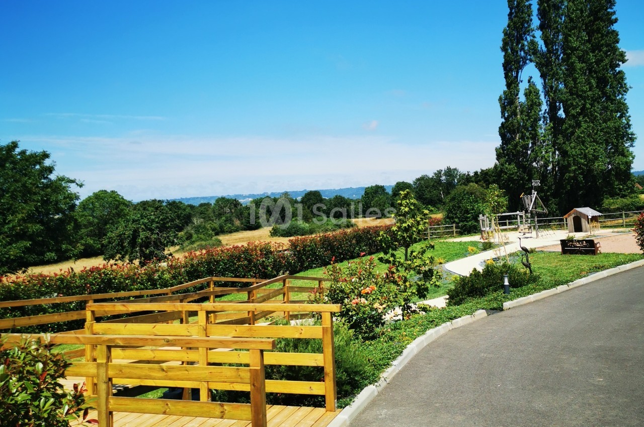 Passerelle en bois surplombant un jardin fleuri avec des arbres et un ciel dégagé en arrière-plan.
