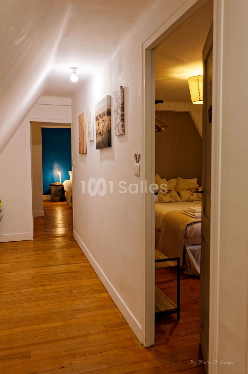 Couloir avec parquet menant à une chambre éclairée, décorée de tableaux et d'une lampe sur table.