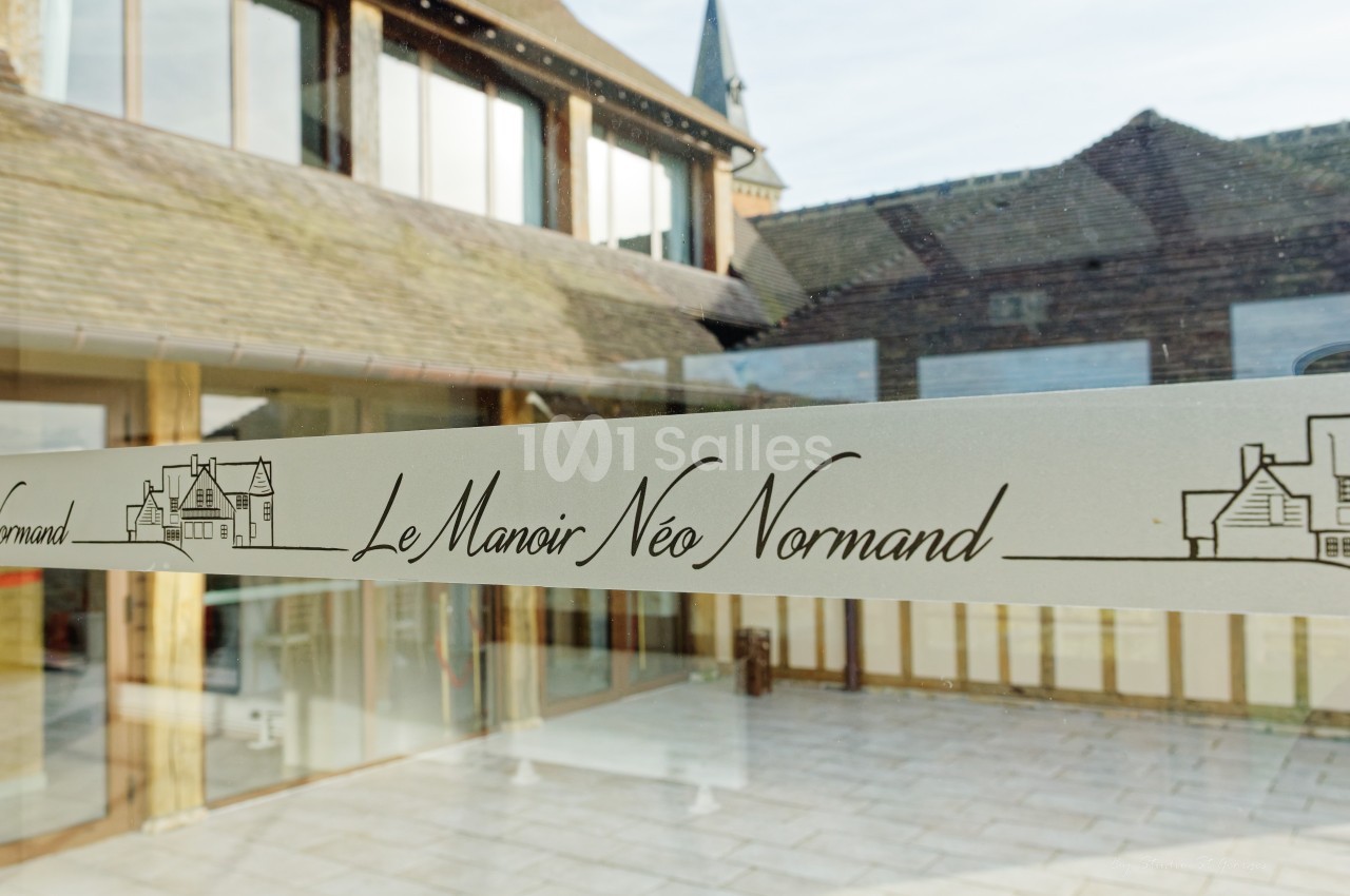 Vue d'une entrée vitrée avec l'inscription ’Le Manoir Néo Normand’ et des bâtiments en arrière-plan.