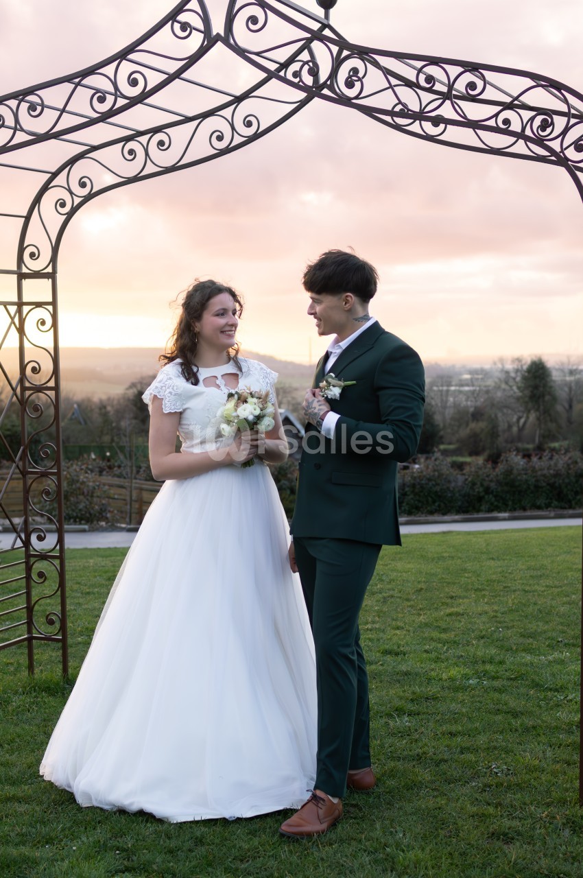 Un couple en tenue de mariage se tient sous une arche métallique dans un jardin au coucher du soleil.