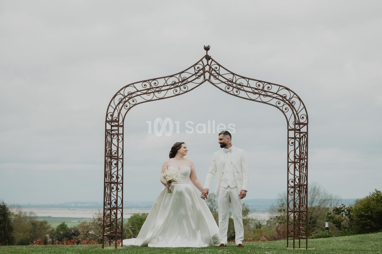 Un couple en tenue de mariage pose sous une arche métallique décorative dans un jardin avec vue dégagée.