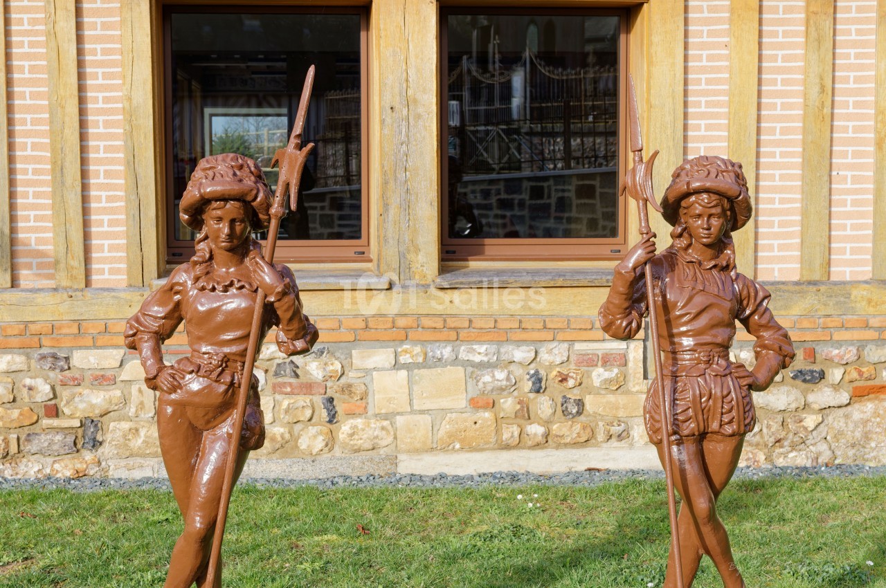 Deux statues en métal représentant des personnages médiévaux devant une façade en briques et pierres avec fenêtres.