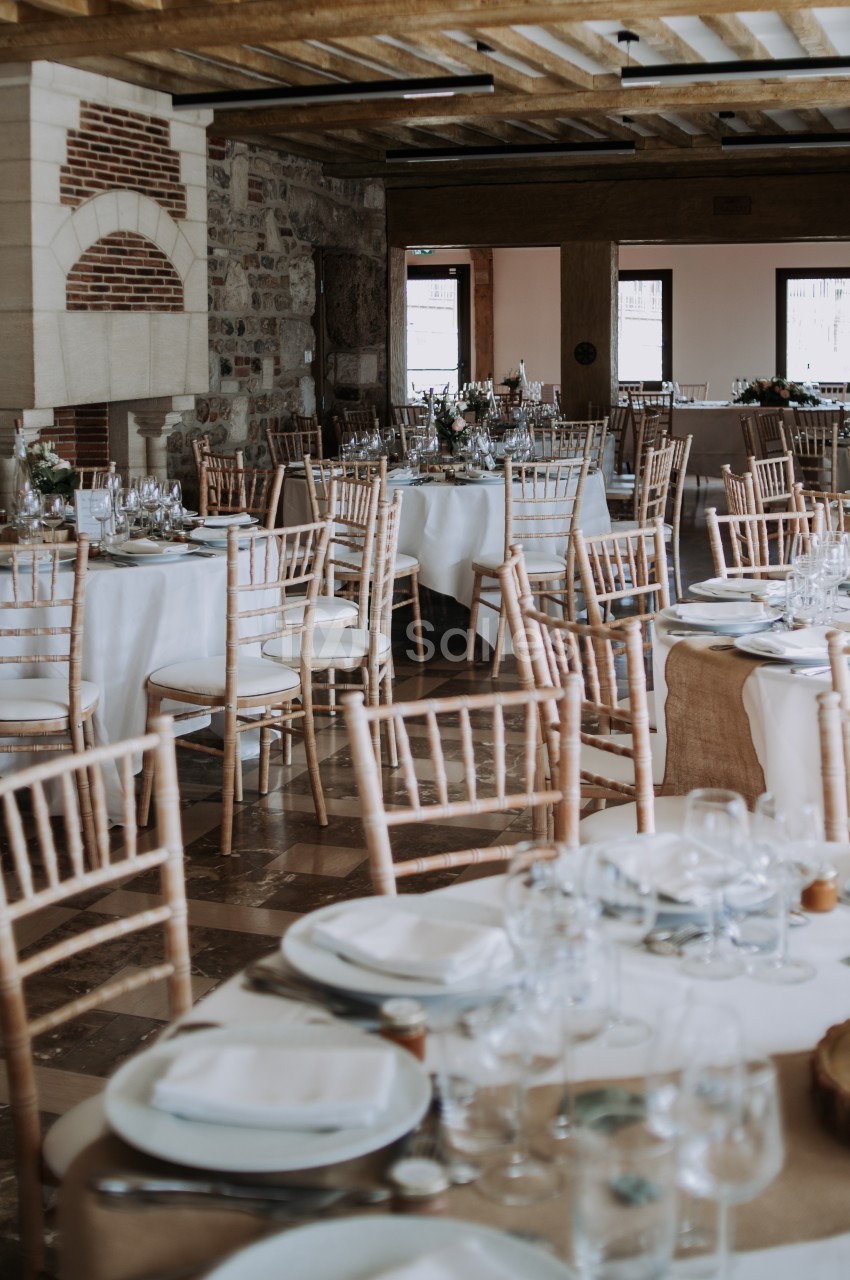 Salle de réception dressée avec tables rondes en blanc, chaises Chiavari bois clair et poutres apparentes.