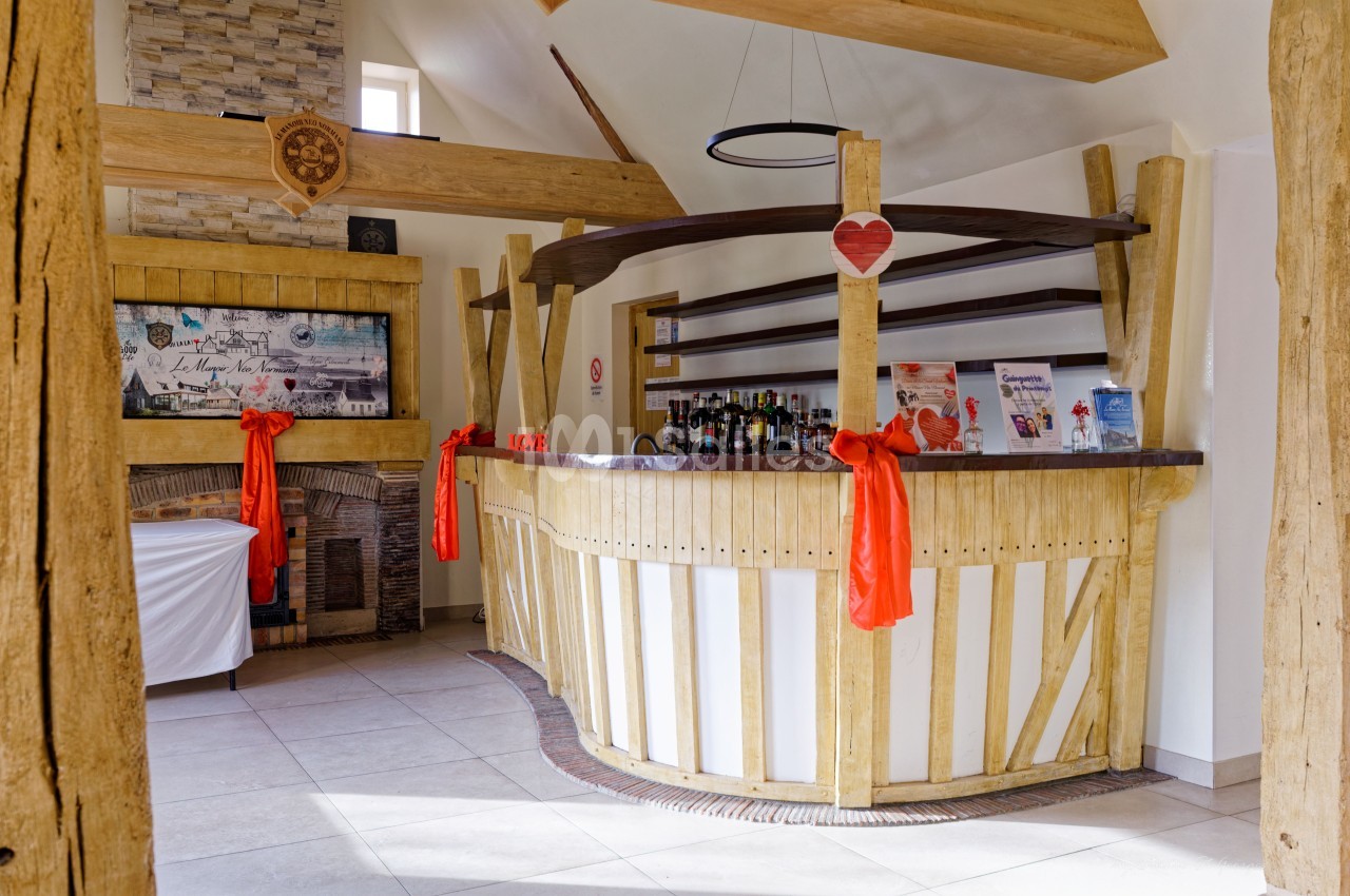 Salle de réception avec bar en bois clair, poutres apparentes, décoration rouge et blanc, bouteilles en arrière-plan.