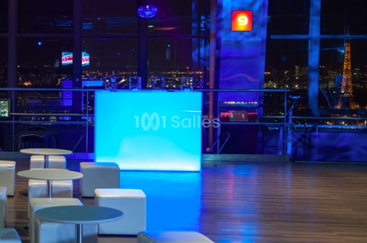 Vue intérieure d'un espace moderne avec un bar lumineux bleu, des sièges blancs et une vue nocturne sur la ville.