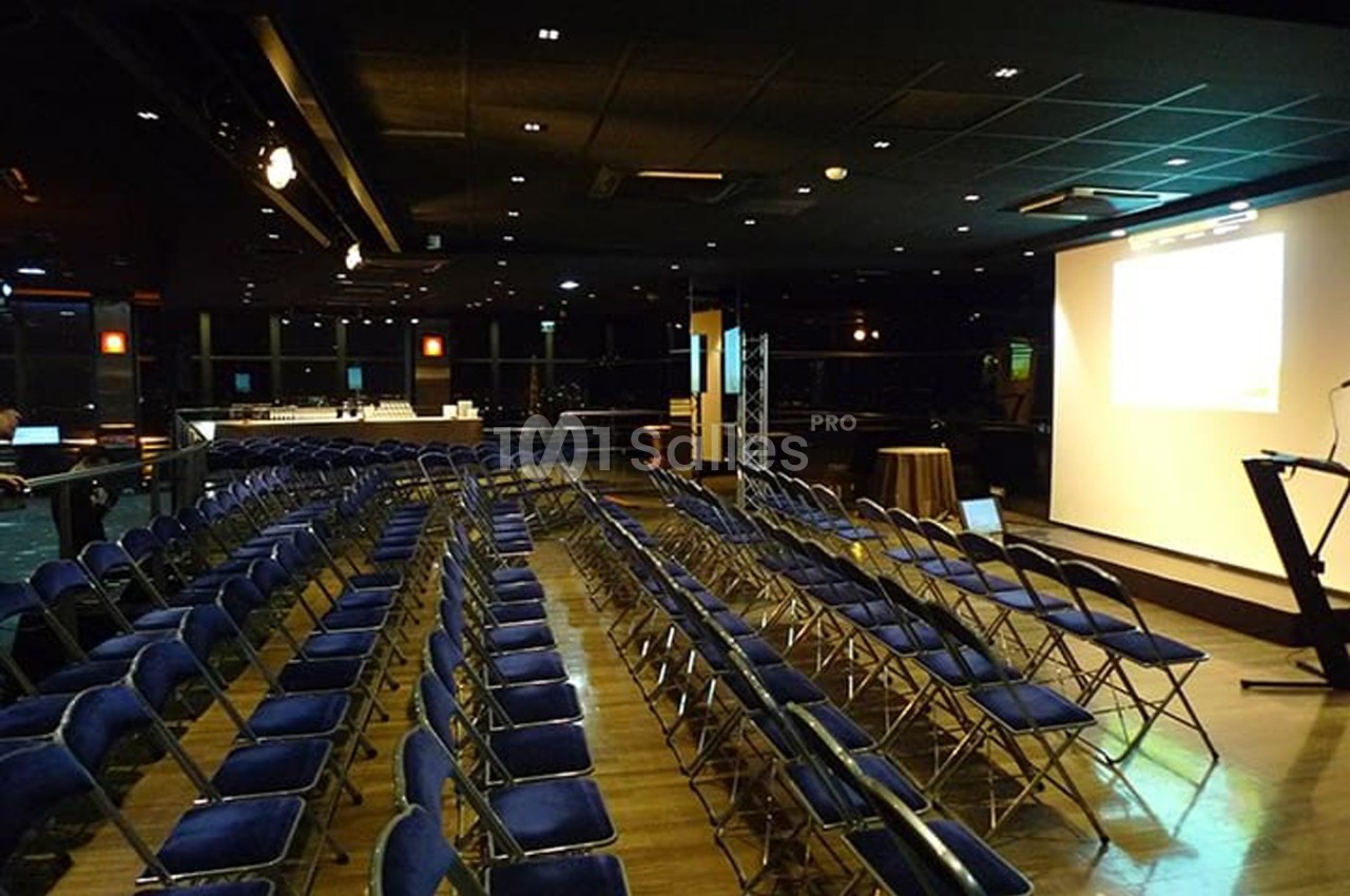Salle de conférence avec des rangées de chaises bleues alignées face à un écran de projection allumé.