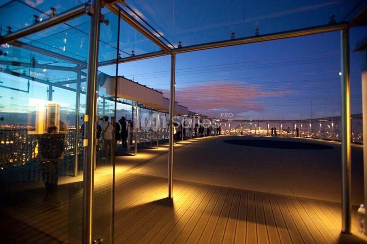 Terrasse panoramique vitrée éclairée au crépuscule, avec des visiteurs observant la vue sur une ville illuminée.