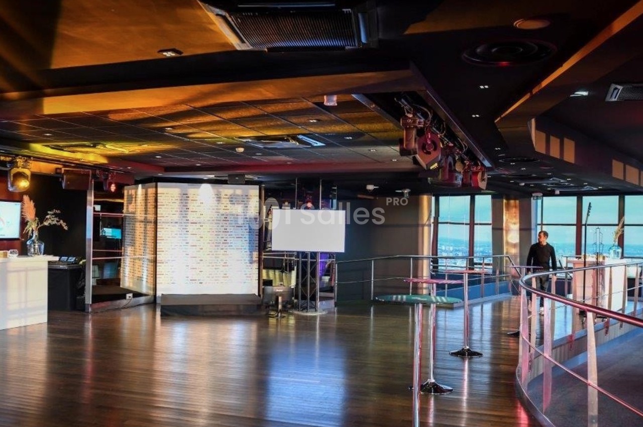Salle spacieuse avec parquet, écrans, bar lumineux et grandes baies vitrées offrant une vue panoramique.